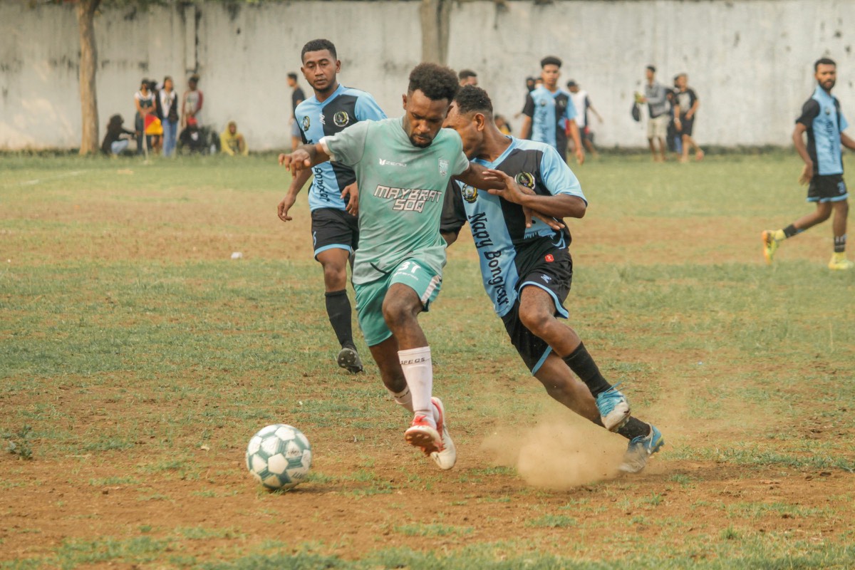 Two players shoulder to shoulder over the ball on a dusty pitch, spectators on the touchline